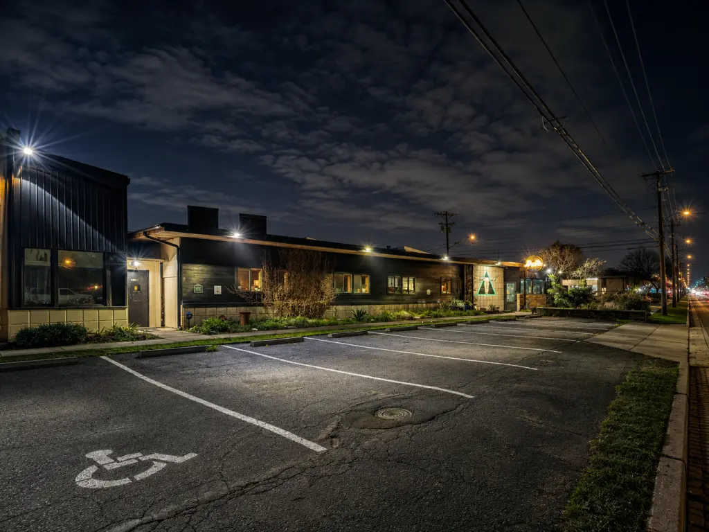 A single-story commercial building features bright exterior security lighting, illuminated windows, and an asphalt parking lot at night.