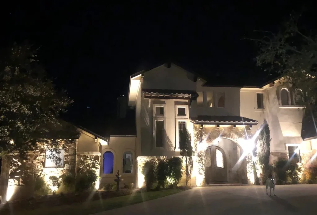 Warm outdoor lighting highlights the stone exterior, arched entryway, trees, shrubs, and architectural features of a luxury estate in Austin, TX.