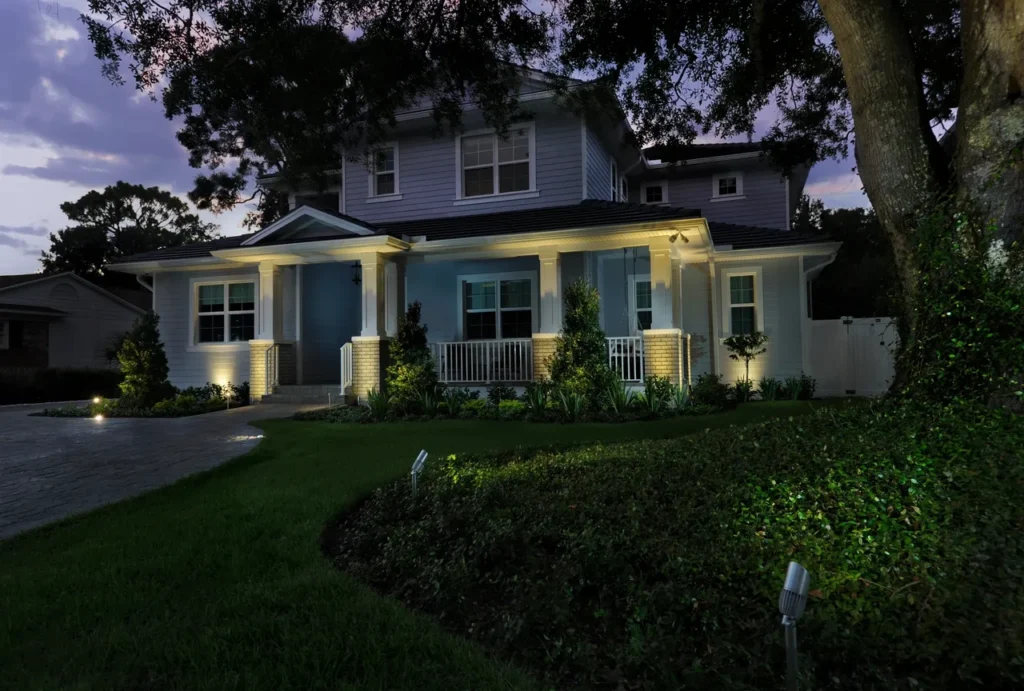 Warm landscape and porch lighting enhances the facade, front yard, flower beds, and curved driveway of a two-story home in Austin, TX.