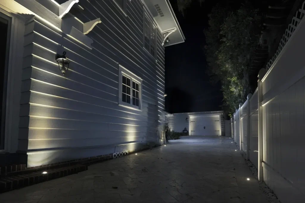 Upgraded architectural landscape lighting featuring recessed ground well lights illuminating the side of a modern white home and driveway.