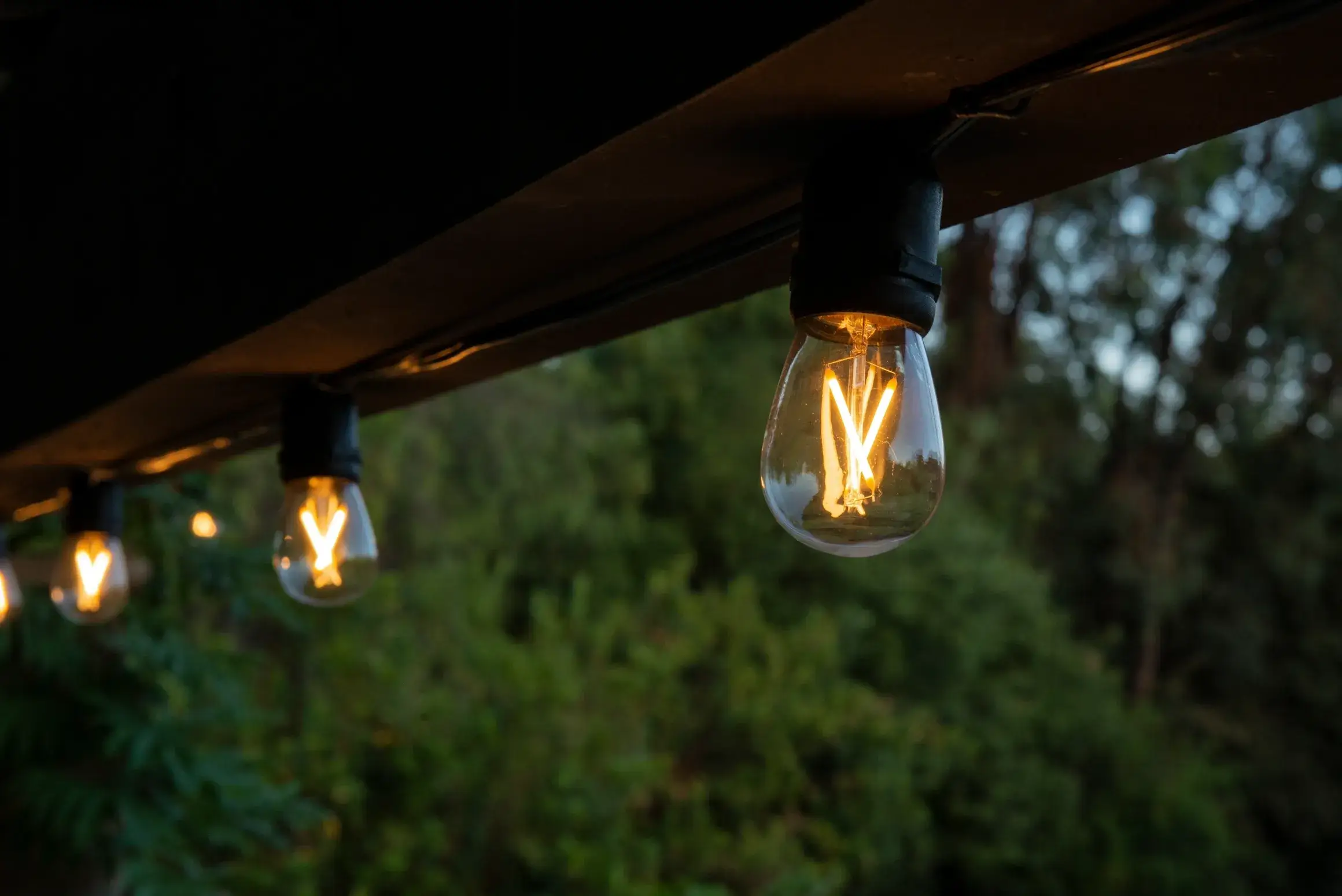 Bistro string lighting by Austin Outdoor Lighting Design creates a warm, inviting glow beneath a patio beam surrounded by greenery.