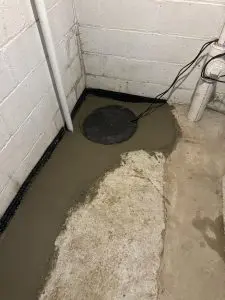 A basement corner with freshly poured concrete, featuring a round cover with attached cables. White pipes run vertically, with a sense of construction progress.