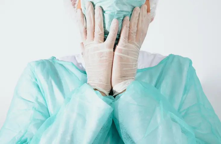 The surgeon holds his head after surgery wearing a green surgical apron and a face mask on a light background.