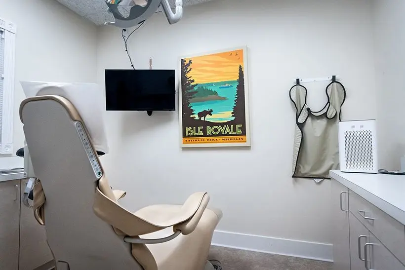 Dental treatment room featuring beige chairs, TV monitor, and wall-mounted protective aprons at Knight Dental Care in Midland, MI