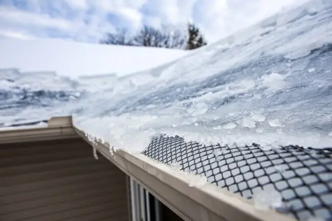 Ice covers the roof of a house, glistening in the sunlight against a clear blue sky.