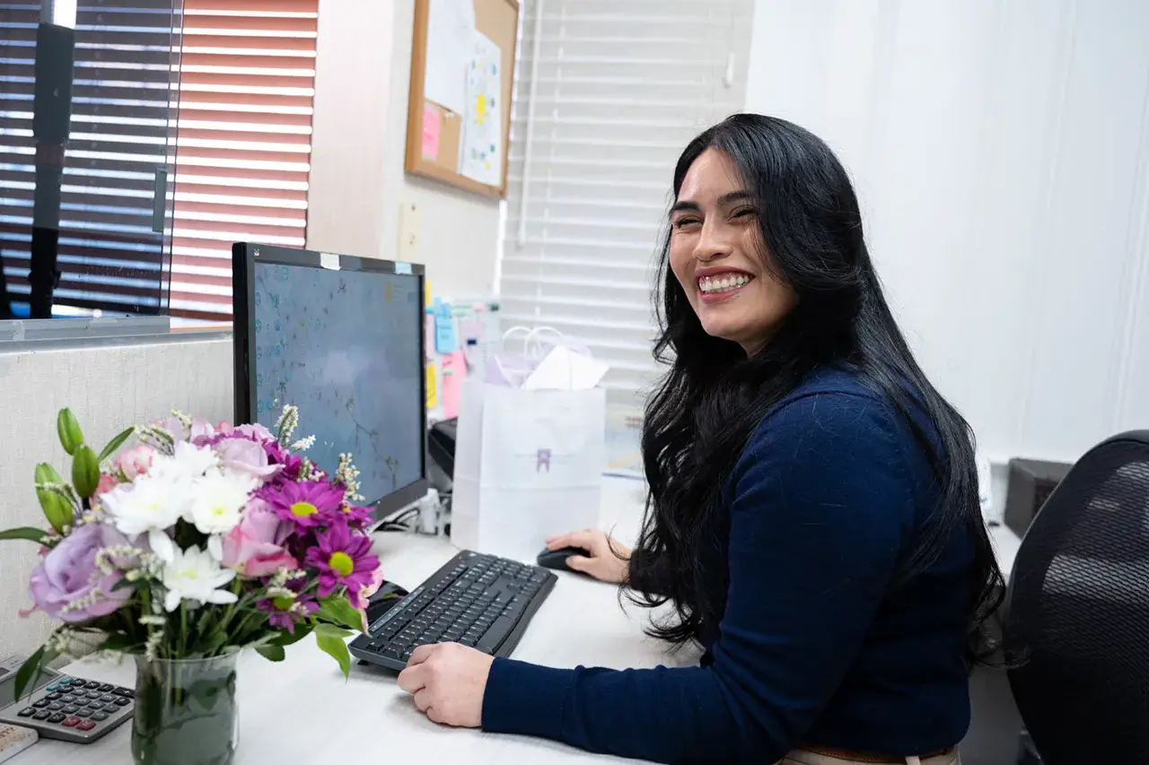 FAQs 2 Receptionist at front desk smiling beside flowers – Dental Office in Westbury, NY – Smallberg & Satornino, DDS, PLLC