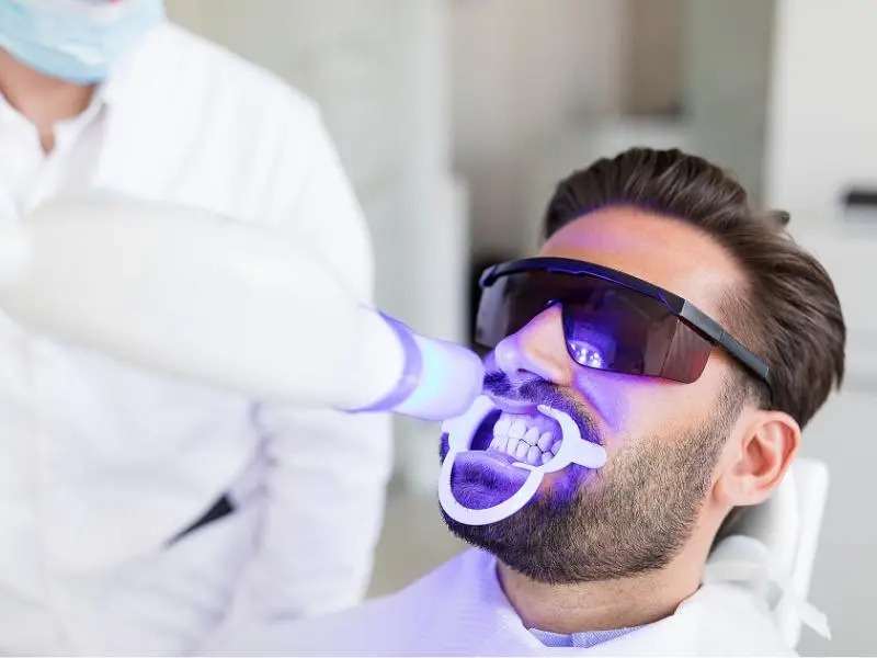 Zoom! Teeth Whitening | VP Dental | Zoom! Teeth Whitening in Raleigh A patient undergoing zoom teeth whitening with a UV light, wearing protective glasses and a cheek retractor