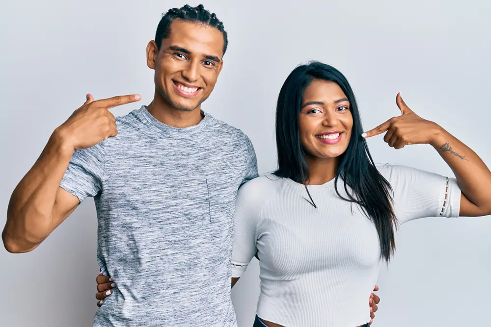 Smile Gallery | VP Dental | Smile Gallery Smiling man and woman pointing at their teeth while standing close together
