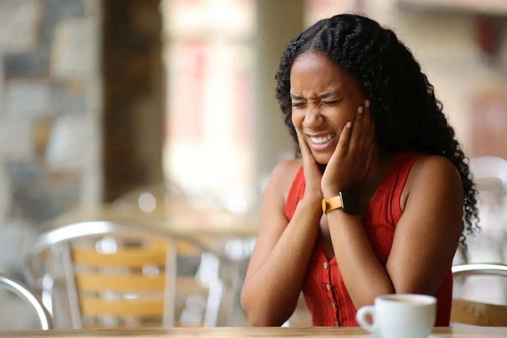 TMJ Treatments | VP Dental | TMJ Treatment in Raleigh Woman in pain, holding her jaw while sitting at a table with a cup of coffee