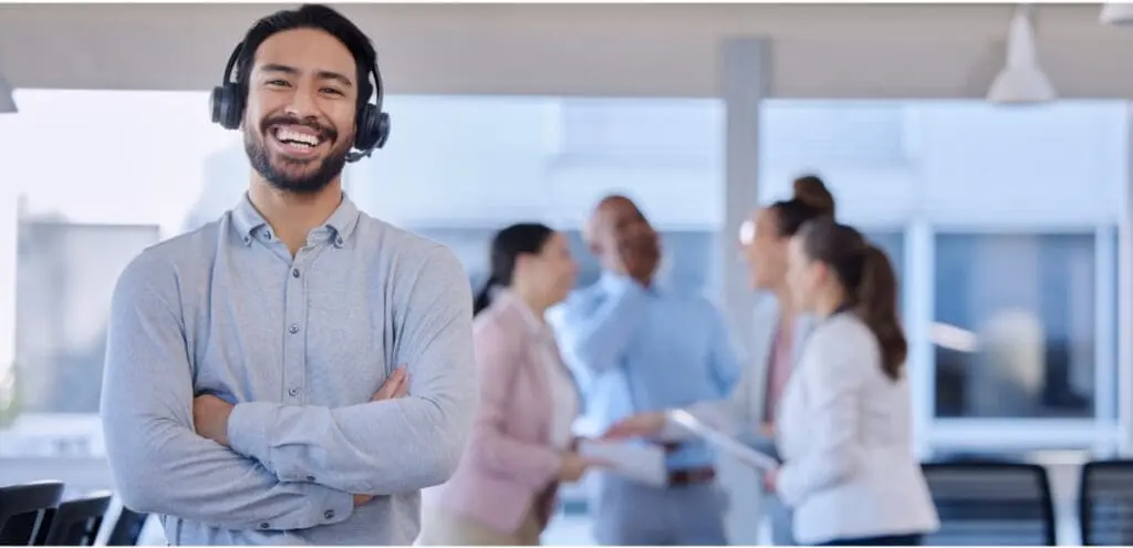 Smiling customer service representative wearing a headset, standing with arms crossed in front of a blurred office background where colleagues are engaged in conversation.