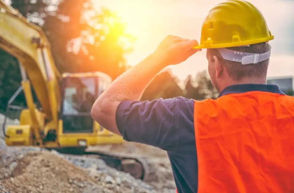 Construction worker wearing a hard hat and an orange safety vest shielding his eyes from the bright sun on a construction site, with heavy machinery in the background.