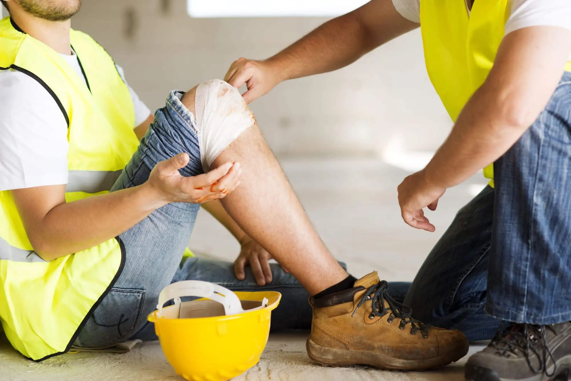 Construction worker sitting on the floor while another person wraps a bandage around his injured knee.