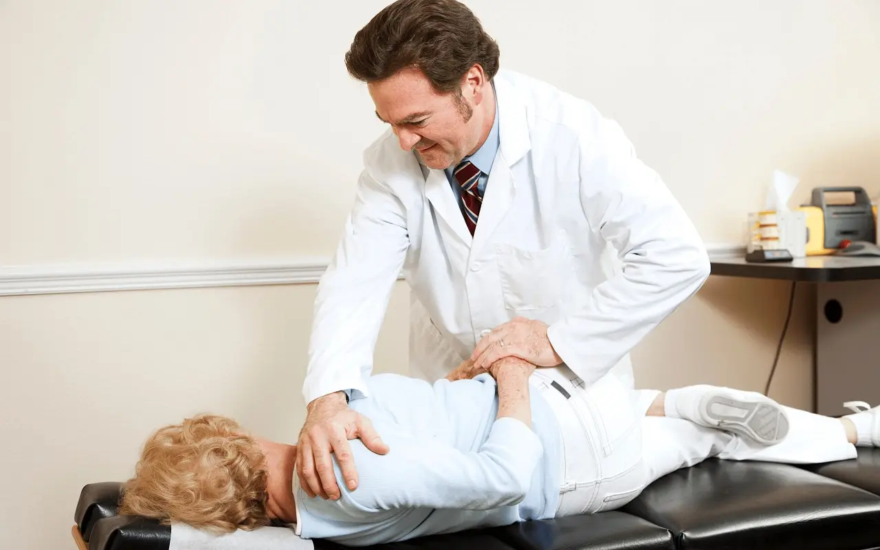 Médico con bata blanca realizando un ajuste o manipulación manual de la columna vertebral en la zona lumbar y la cadera de una paciente, mientras esta se encuentra tumbada en la camilla de tratamiento para aliviar de forma no invasiva el dolor de espalda relacionado con el trabajo.