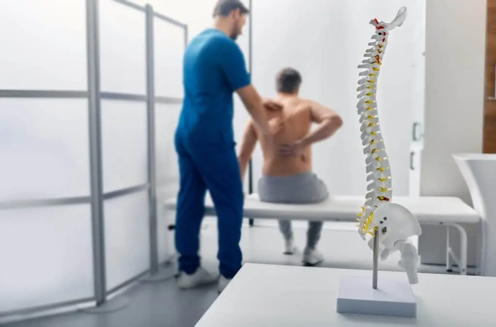 Médico o fisioterapeuta que examina el dolor lumbar de un paciente varón con ayuda de un modelo anatómico de la columna vertebral