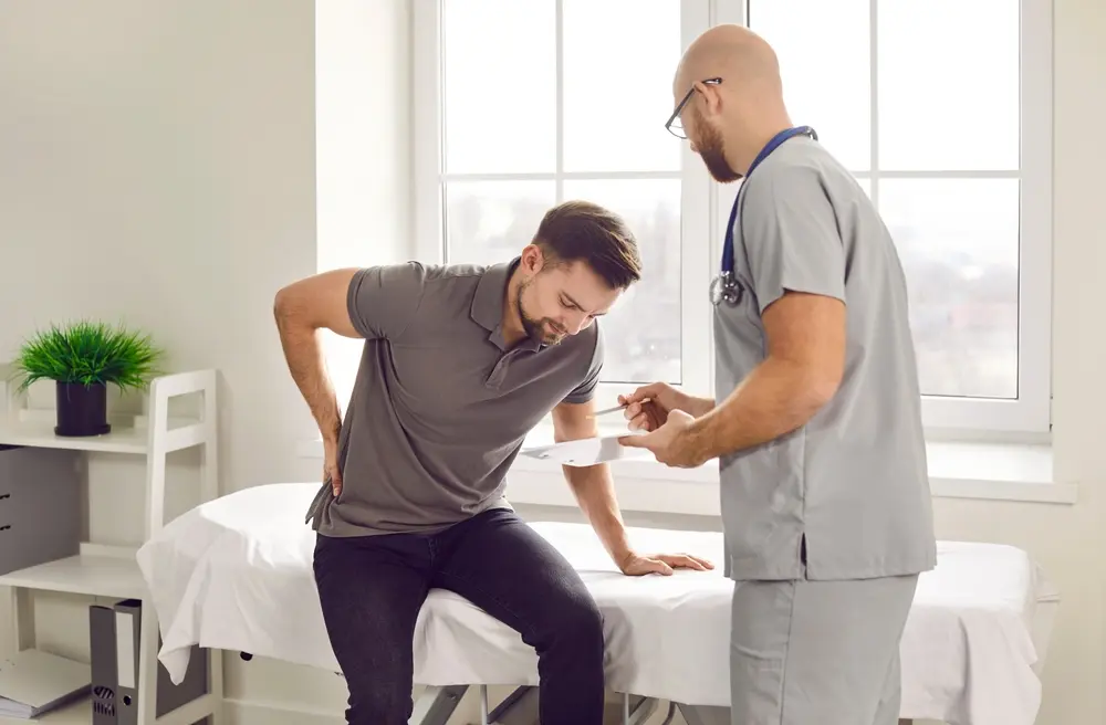 Adult patient sitting and holding lower back in pain while a spine specialist stands in front asking about symptoms during consultation.