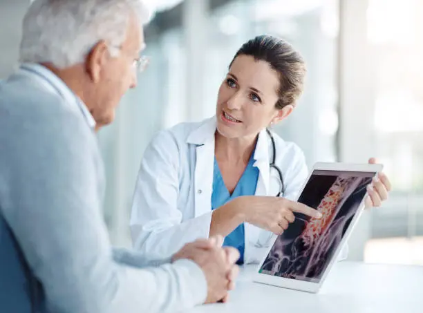 Young female spine specialist pointing to an area on a spine X-ray while explaining symptoms to an elderly patient.