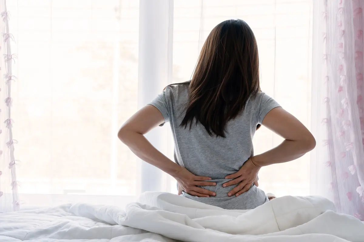 Triste joven asiática tocando espalda sintiendo dolor de espalda malestar matutino dolor lumbar bajo riñón muscular sentarse en la cama después de mal sueño despertar en colchón incómodo flexión. Concepto de mujer estirándose sufriendo dolor de espalda repentino.