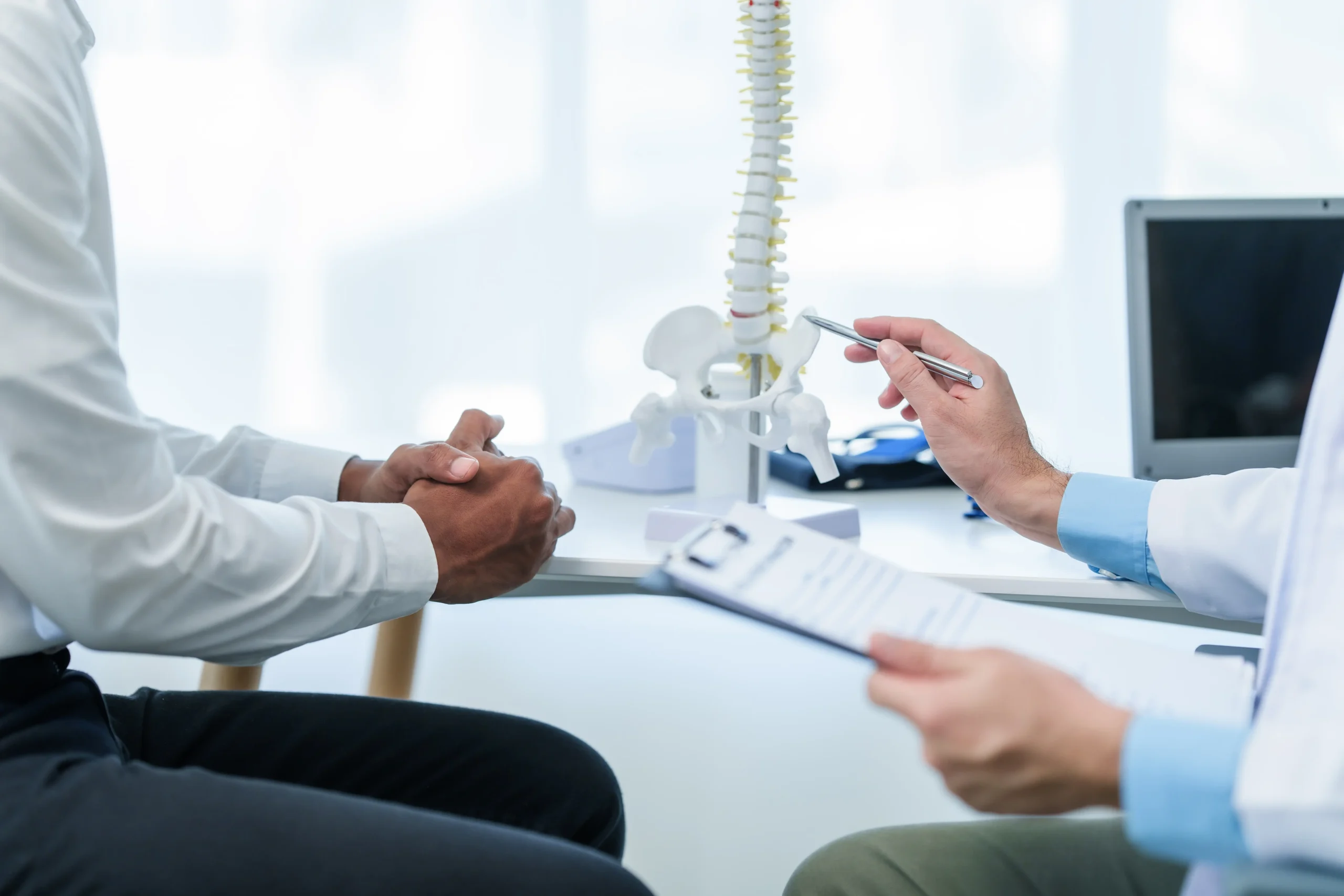 Doctor showing spine model to patient while explaining causes of musculoskeletal pain and treatment options in a medical office.