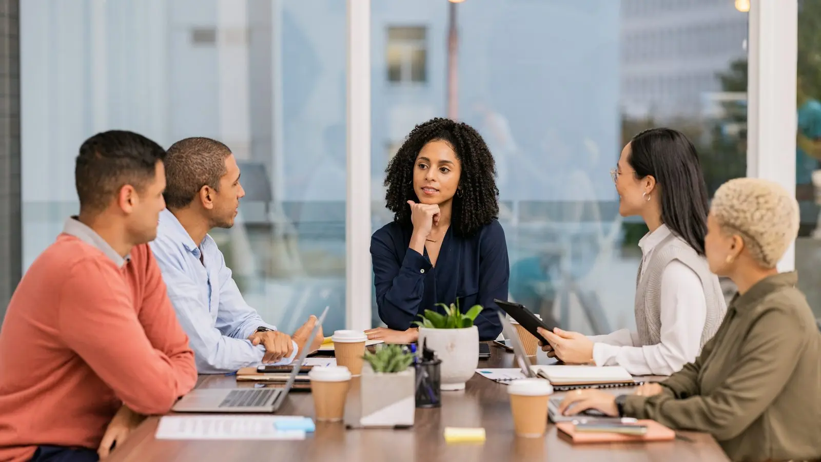 A home services owner, office manager, operations manager, and marketing coordinator lean over a single sheet of paper at a conference table in a contractor office, a job schedule board out of focus behind them.