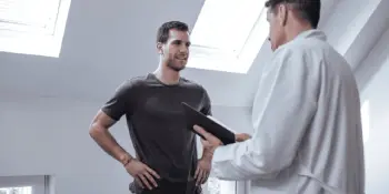 Confident doctor in white coat discussing treatment with fit male patient holding clipboard in bright medical exam room for car accident or workers’ comp recovery