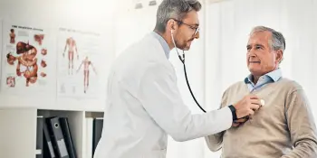 Doctor using stethoscope to examine senior male patient in medical office, checking for injuries from car accident or workers’ compensation claim
