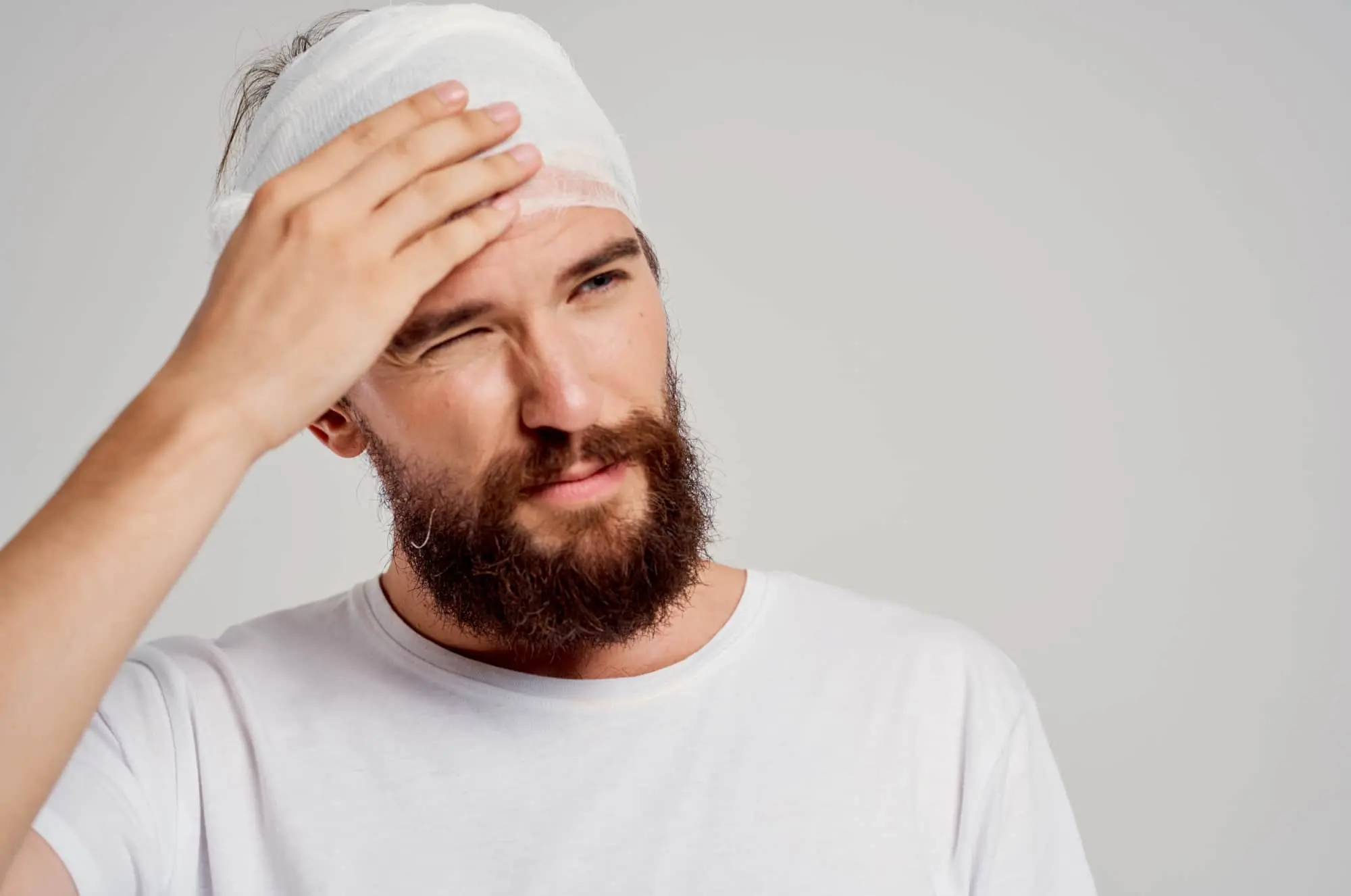 Bearded man with head bandage holding forehead in pain, showing signs of headache or concussion after accident or injury