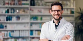 Smiling male doctor in white coat with glasses standing confidently in front of pharmacy shelves, ready to treat car accident and workers’ comp injuries