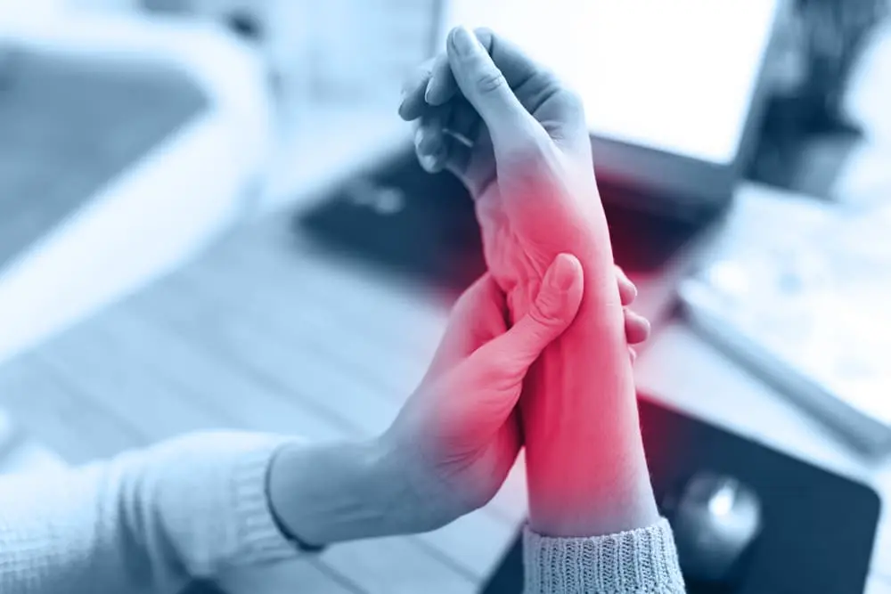 Mujer sujetándose la muñeca dolorida con inflamación roja destacada mientras trabaja con el ordenador portátil: síntoma de lesión por esfuerzo repetitivo.