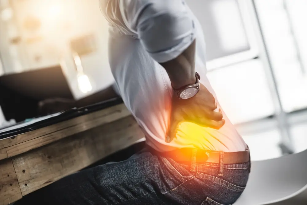 Un hombre sentado en una oficina sujetándose la parte inferior de la espalda con dolor, mostrando molestias en la columna vertebral y lesiones de espalda en el lugar de trabajo.