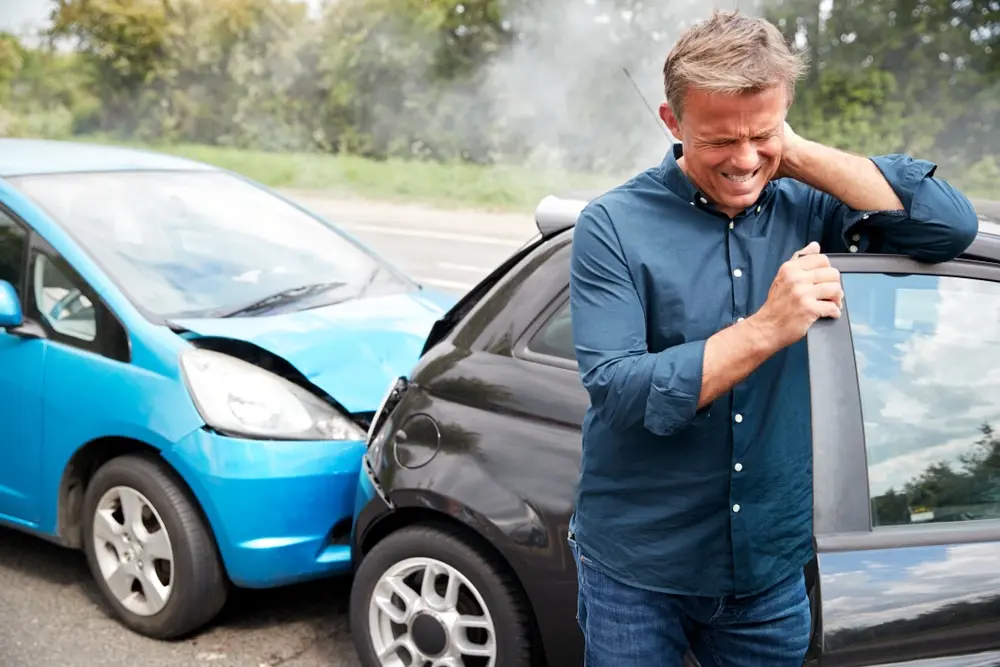 Hombre de mediana edad que se sujeta el cuello con un dolor intenso, de pie junto a unos coches azules y negros dañados tras una colisión en la carretera, y que presenta los síntomas habituales de latigazo cervical o distensión cervical tras un accidente de tráfico.