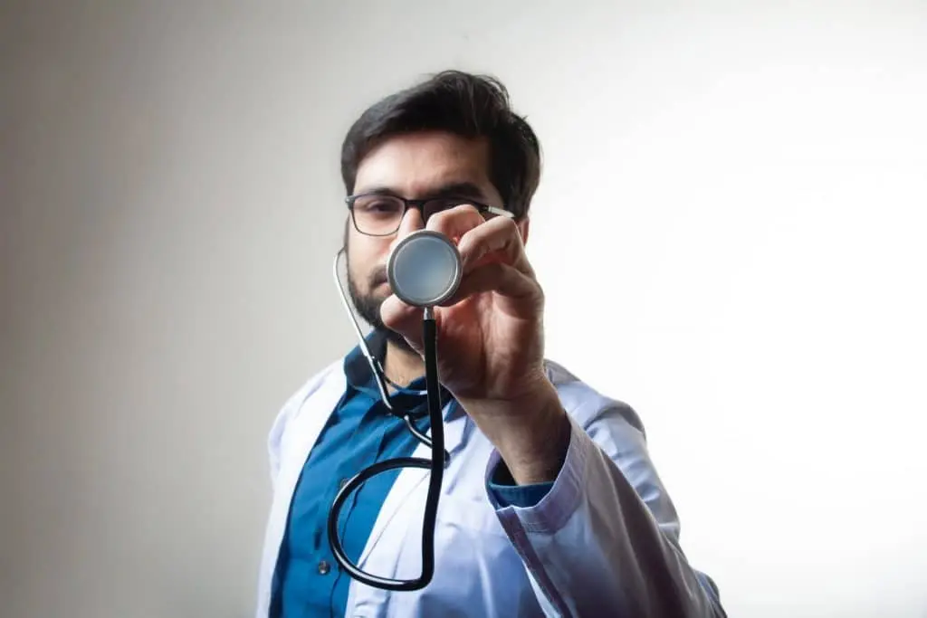 Male doctor in blue scrubs and white coat holding stethoscope up to camera in medical office, ready to examine patient after car accident, workplace injury, or slip and fall in New York