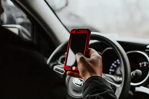 Hombre que sostiene un smartphone rojo en una mano mientras sujeta el volante dentro de un coche, lo que ilustra la conducción distraída y el mayor riesgo de accidentes de tráfico o lesiones.