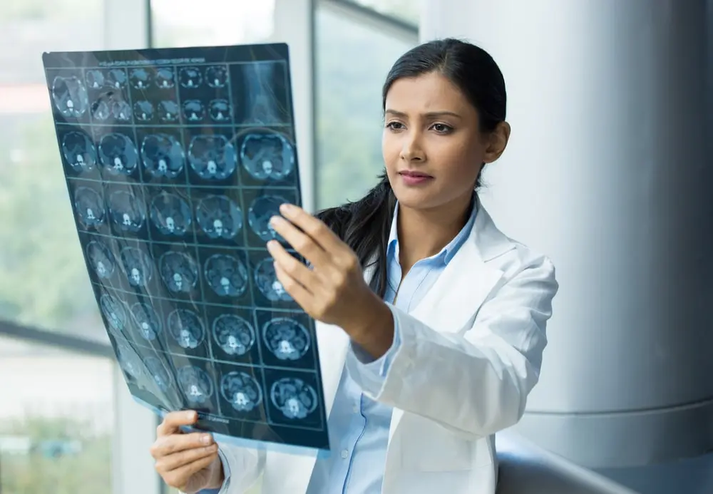 Una neuróloga o radióloga con bata blanca examina una radiografía de resonancia magnética cerebral en una mesa de luz en un hospital o una clínica, evaluando un traumatismo craneal o una lesión neurológica provocada por un accidente de tráfico en Nueva York.