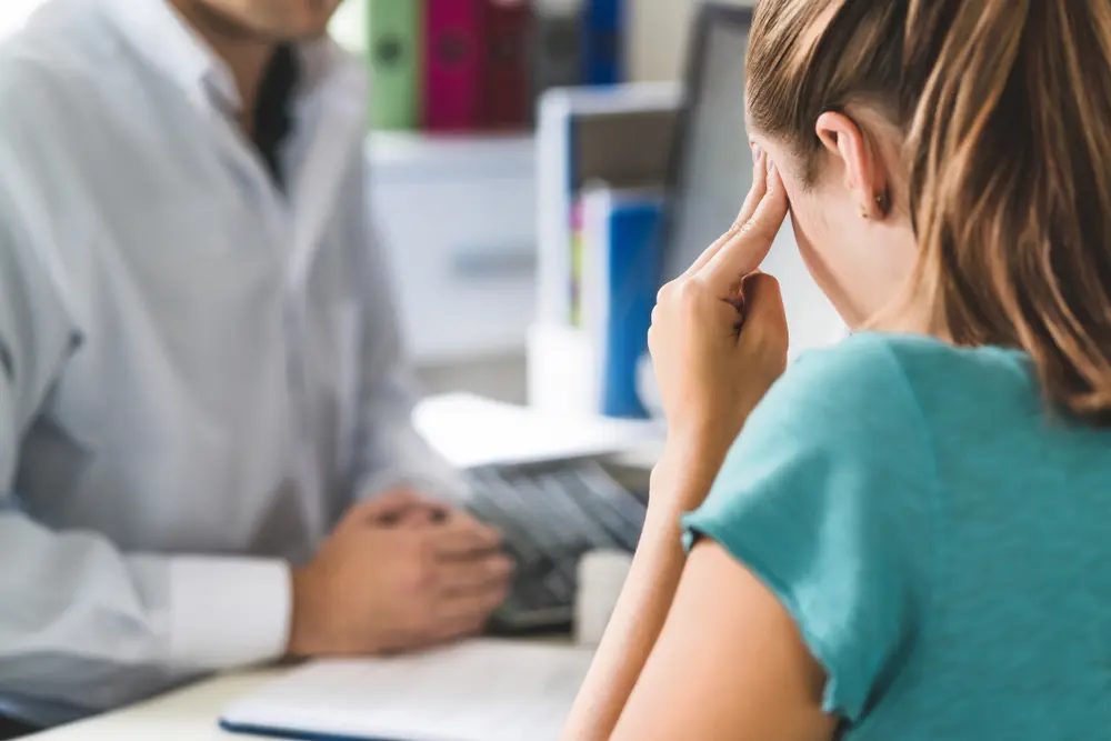 A young woman with head pain sits in front of a doctor, holding her forehead as she experiences post-accident brain trauma symptoms.