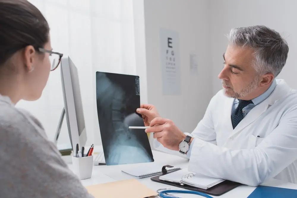 Male doctor in white coat pointing with pen at lumbar spine X-ray film on lightbox while consulting female patient in medical office, discussing back injury diagnosis after accident or workplace strain