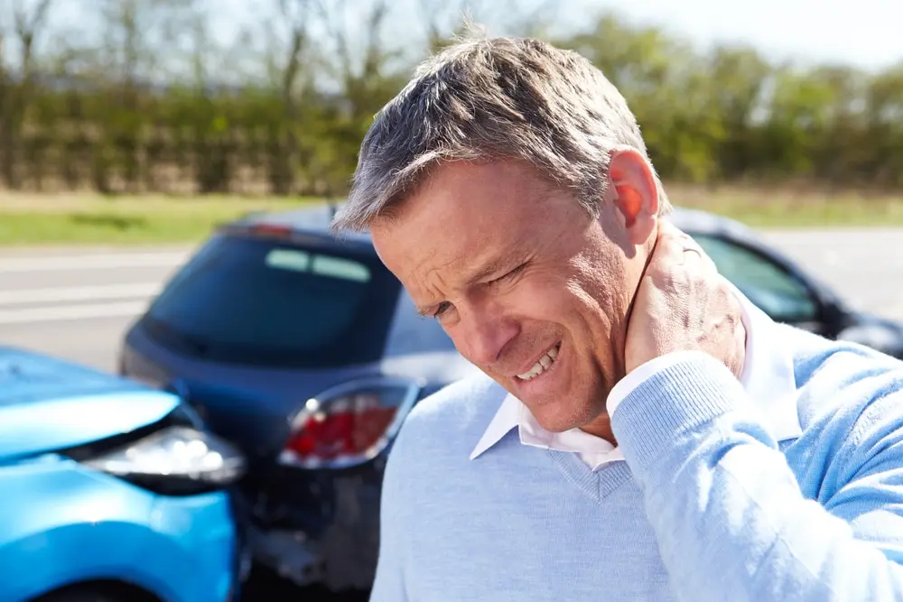 Man grimacing in neck pain after a car accident, holding his neck while standing near two damaged vehicles on the road.