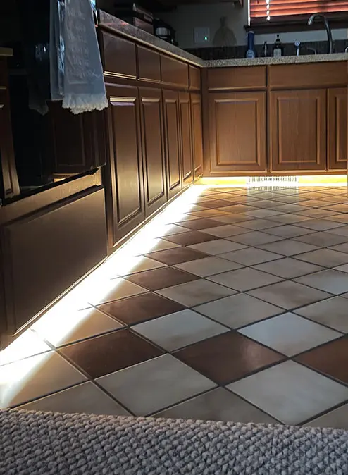 Warm under-cabinet lighting spilling onto a checkered tile kitchen floor, highlighting wood cabinetry and creating a cozy evening ambiance.
