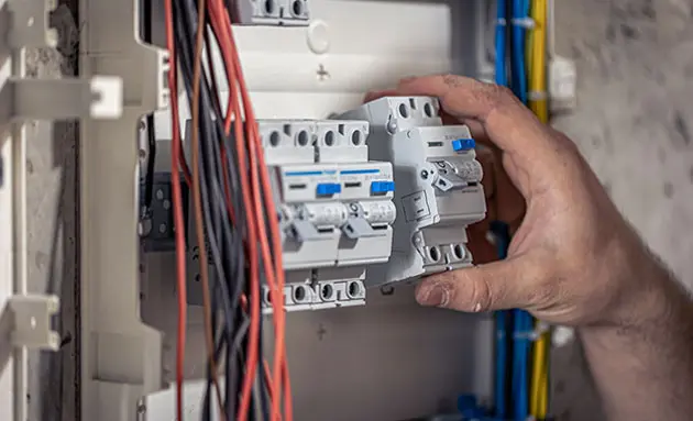 Close-up of an electrician's hand fitting circuit breakers into an open electrical panel with red, black, and blue wiring.