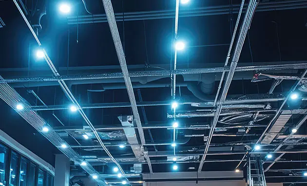 Rows of bright LED high-bay lights on an exposed industrial ceiling with metal cable trays, conduit, and ductwork throughout the space.
