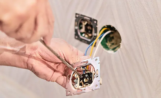 Close-up of an electrician's hands using a screwdriver to connect color-coded wires to an electrical outlet mounting plate.