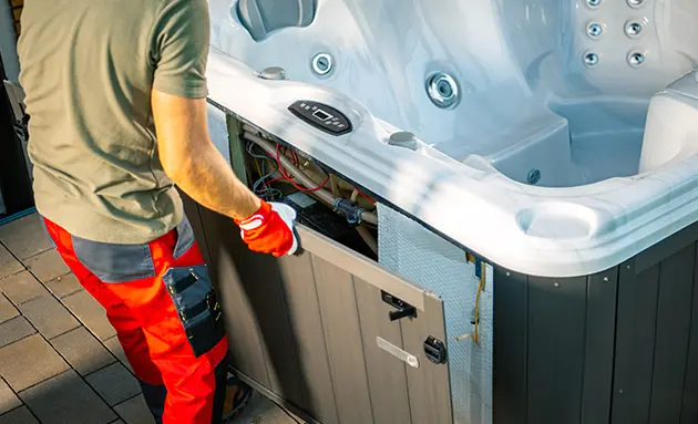 Technician in work clothes and gloves connecting the electrical wiring of a hot tub with the side access panel removed.