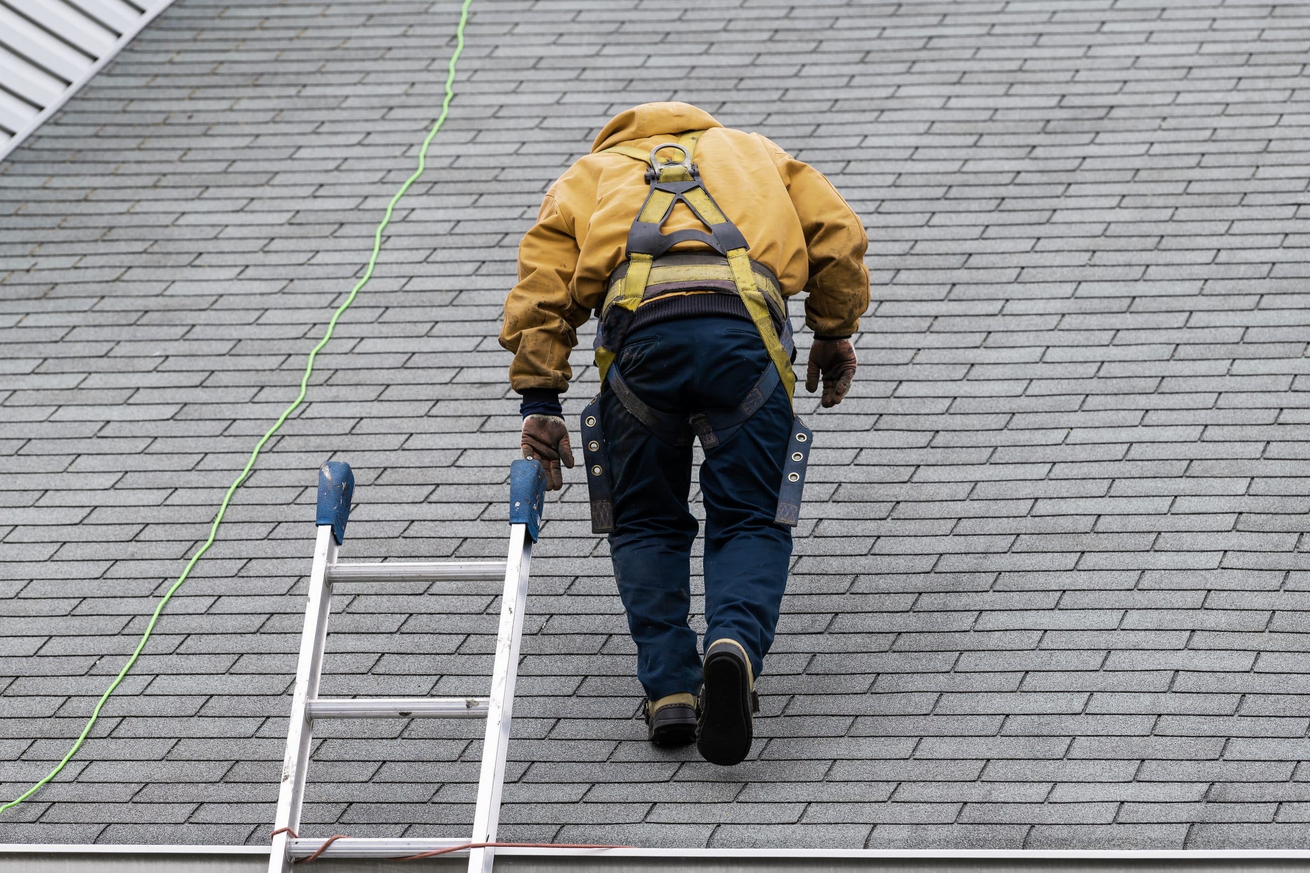 House during day with gray color Single Family Home and construction man in yellow uniform walking on roof shingles and ladder during repair