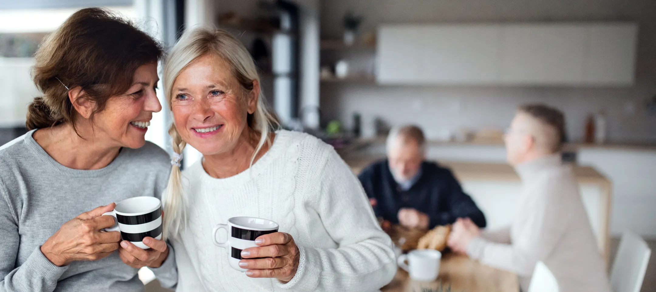 two middle aged women in kitchen drinking coffee with husbands in the background