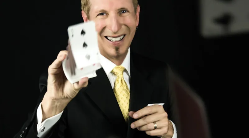 Magician performing a card trick with a smile, showcasing a deck of cards to the audience.