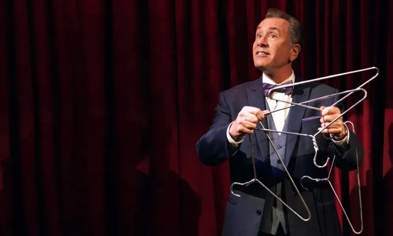 Magician performing a comedy magic trick with a metal hanger, smiling and engaging with the audience.