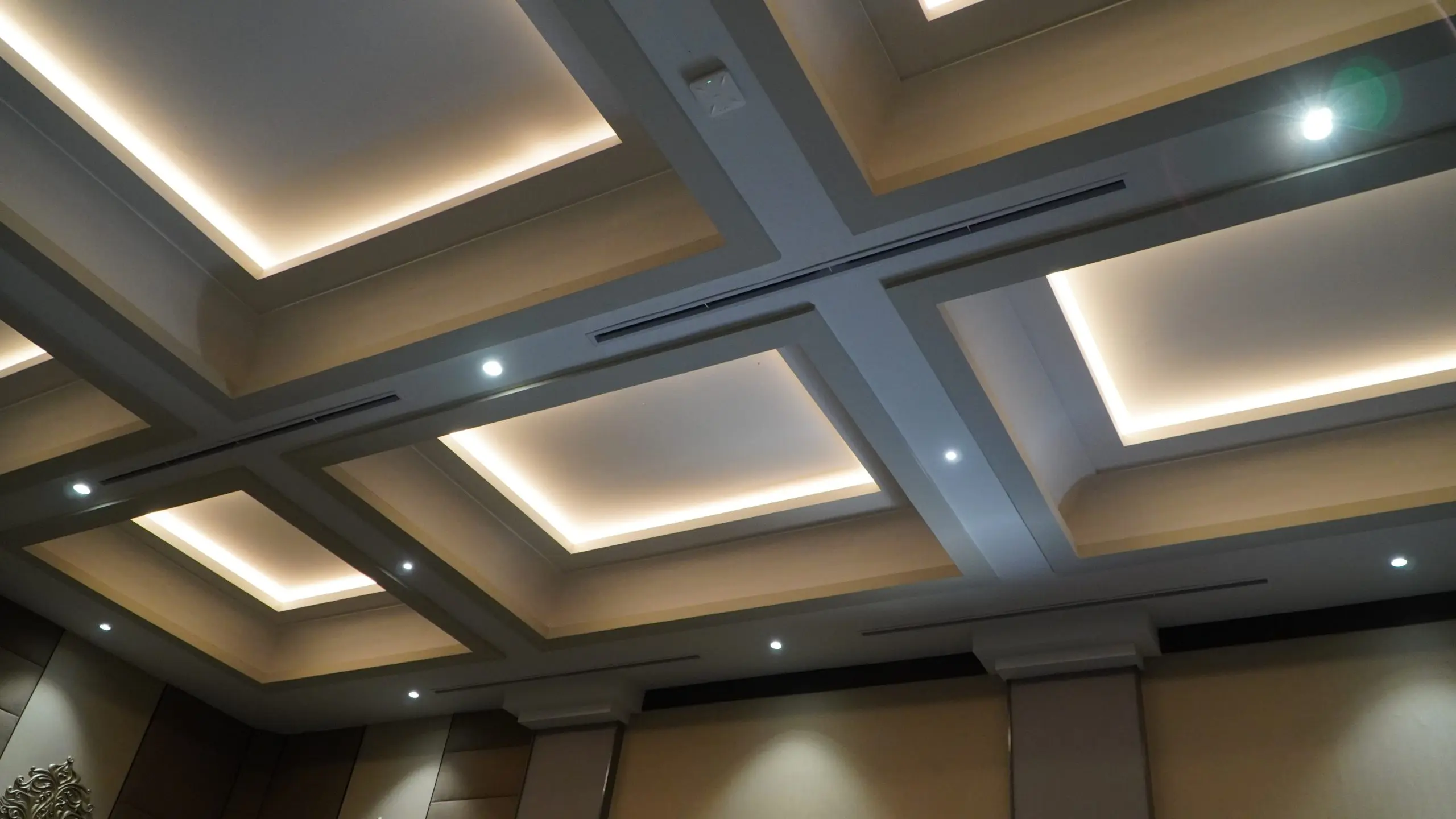 A wide-angle, upward-looking shot of a modern, coffered ceiling design featuring recessed square panels with warm LED cove lighting and integrated recessed spotlight fixtures.
