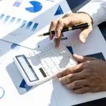 Manos utilizando una calculadora y un bolígrafo en documentos financieros con gráficos, reflejando los servicios de revisión de pólizas de seguros y evaluación financiera.
