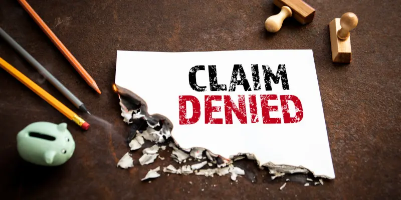 Claim denied notice on torn paper, surrounded by pencils, a piggy bank, and wooden sellos, symbolizing denied home insurance claims and the need for public adapting assistance.