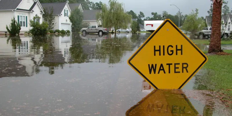 Zona residencial inundada en League City, Texas, con un prominente signo de advertencia "HIGH WATER", que ilustra el impacto de los daños causados por inundaciones relevantes para las reclamaciones de seguros.