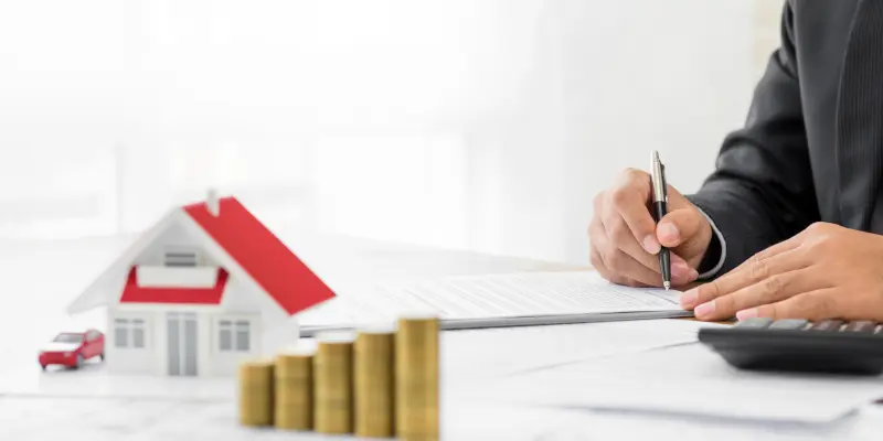 Persona firma documentos con una casa modelo y pilas de monedas, representando servicios de evaluación de seguros por los Ajustadores Públicos de Santex.